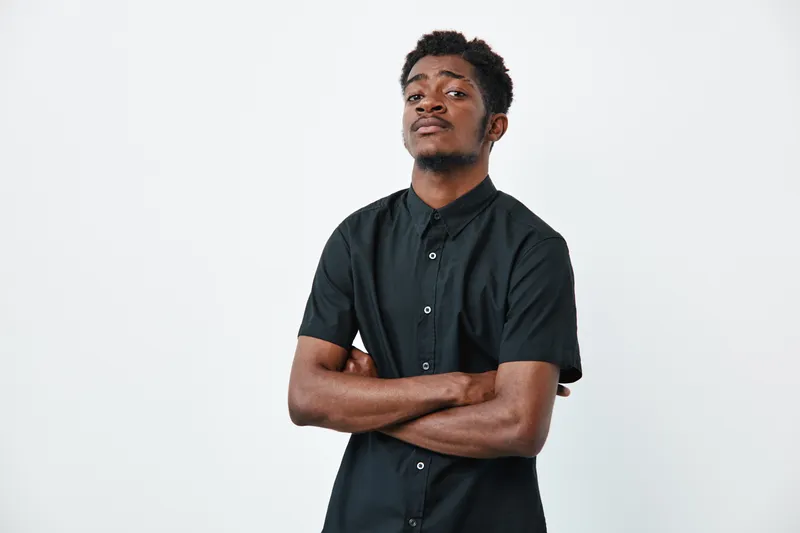 Young black man with dark skin wearing black short sleeve shirt standing with crossed arms looking confident on white background. Portrait of African ethnicity male model.