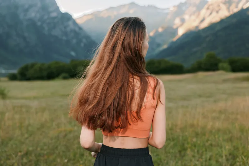 A young woman stands in a sunlit meadow, facing away from the camera. She wears an orange crop top and dark bottoms, with distant mountains behind, inviting a sense of freedom, realism, and natural