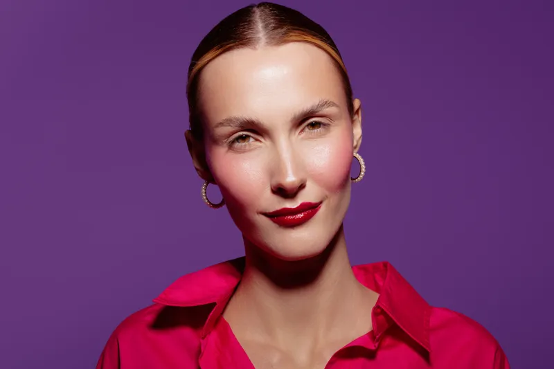 Portrait of confident woman with red lipstick and hoop earrings wearing bright pink shirt against purple background. Concept of beauty, fashion, and modern style with studio lighting.