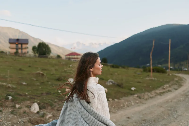 A reflective woman in a cozy sweater stands outdoors in a mountain landscape, gazing into the distance. A moment of solitude and calm, nature inspired lifestyle.