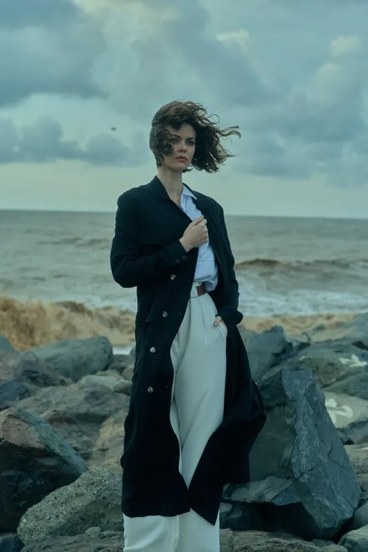 Fashion model on the coastline wearing a long black coat over white trousers, wind swept hair, confident pose by rugged rocks and sea