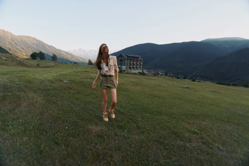 A young woman with long hair walks across a grassy field near a mountain lodge during late afternoon, enjoying a peaceful moment in nature.
