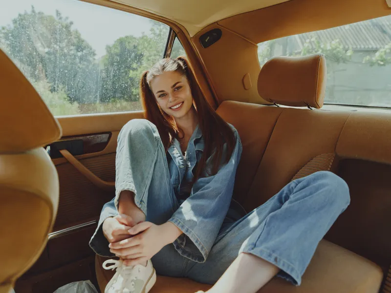 Smiling young woman in casual denim outfit sitting in a car, enjoying a sunny day. Perfect for travel and lifestyle themes.
