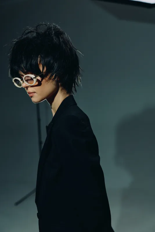 Stylish woman with short black hair wearing white round glasses and black blazer against dark background.