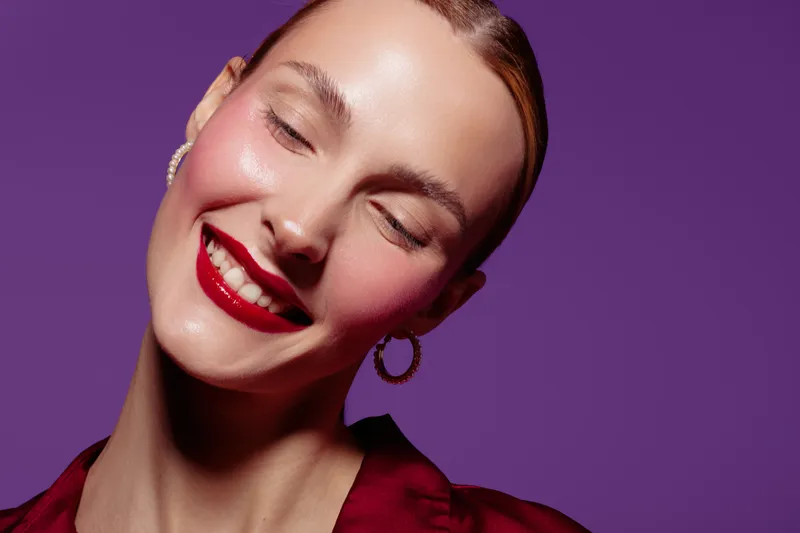 Smiling woman with red lipstick and hoop earrings poses with closed eyes against purple background. Concept of beauty, makeup, fashion, and confident female portrait in studio lighting.