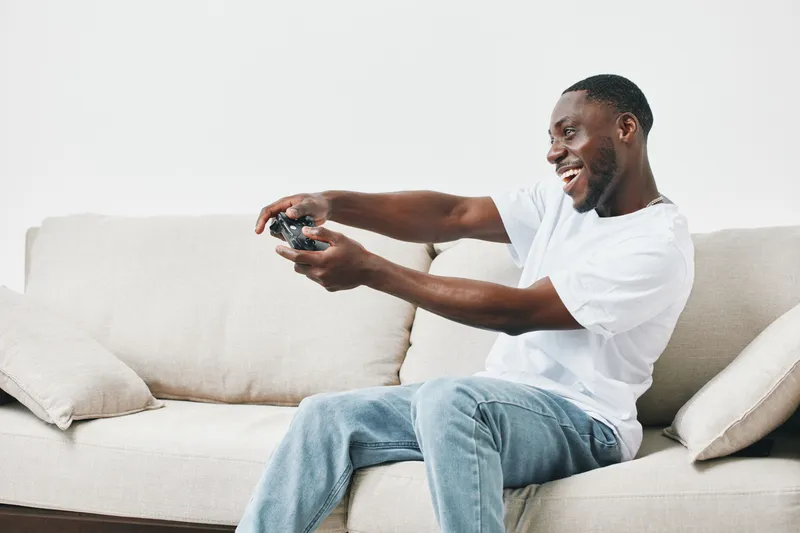 Happy young African man enjoying gaming at home on a sofa, wearing casual clothing, immersed in his favorite video games, creating a cheerful atmosphere