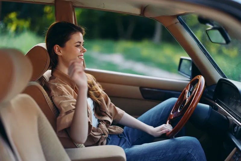 Young woman smiling inside a vintage car, wearing casual attire, enjoying a sunny day in nature, evoking feelings of freedom and adventure.