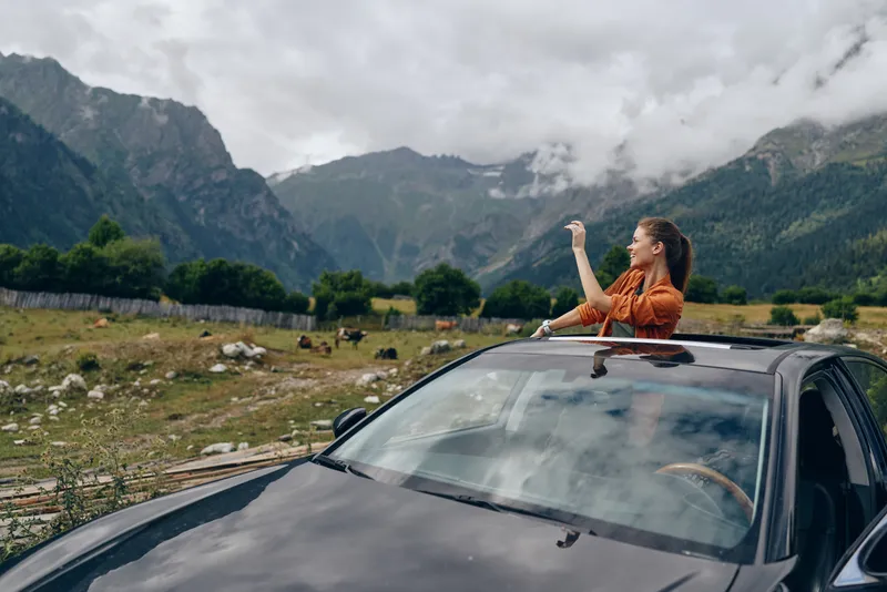Joyful woman sticks her arm from a sunroof during a road trip in a mountainous landscape, smiling and energetic as the car cruises through open countryside.