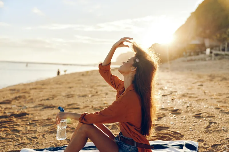 Woman on beach at sunset, sunlight around her holding water bottle, focusing on hydration, summer outdoors wellness and relaxation, sandy coast with ocean horizon, soft golden light