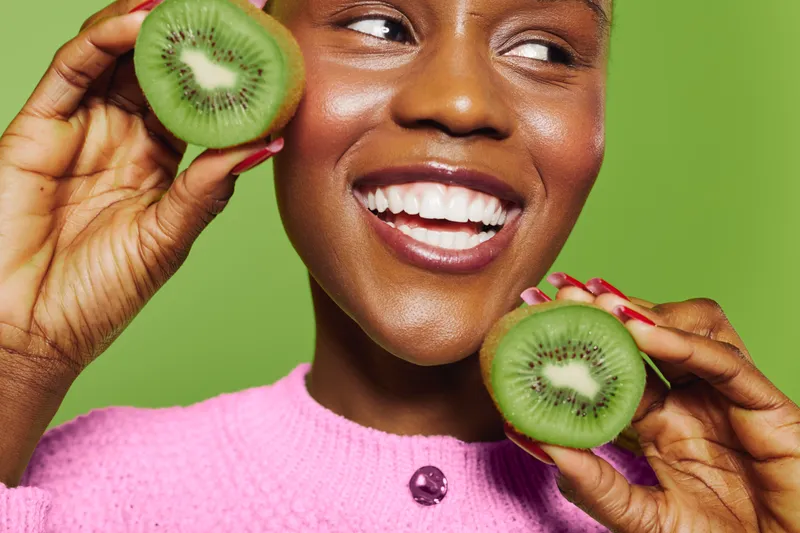 Smiling happy black woman holds two halves of fresh kiwi near face on green background. Concept of healthy lifestyle, natural food, fruit, and positive mood in beauty and wellness.