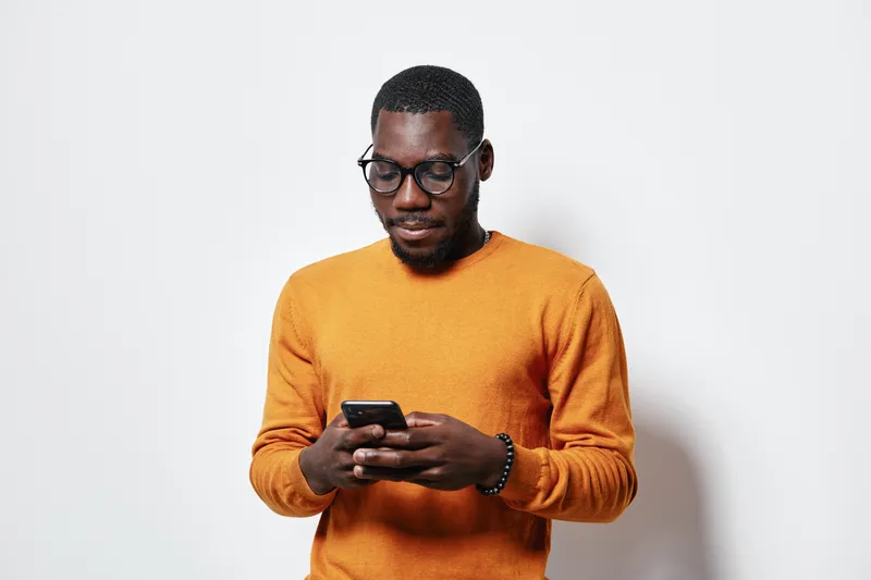 African man with dark skin wearing glasses and orange sweater using smartphone for texting or browsing on white background. Casual adult male focused on mobile device communication.