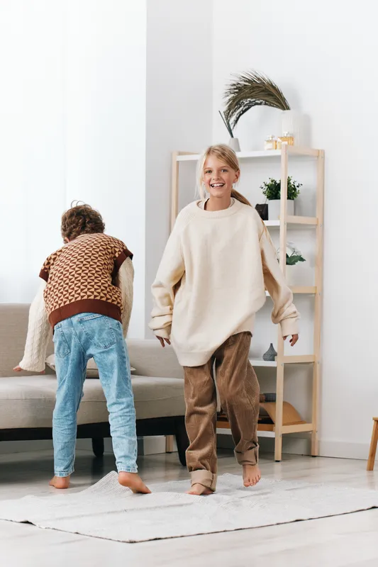 Authentic scene of children at home engaged in playful activity, cozy outfits, simple living room setting, genuine smiles, everyday life in a light, relaxed moment.