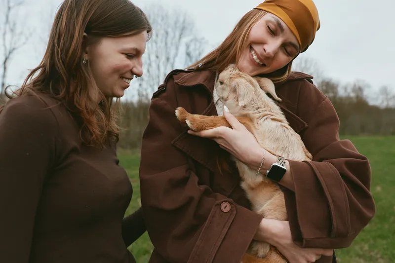 Two young women enjoy a tender moment outdoors as one woman in a brown coat lovingly holds a baby goat while the other smiles warmly beside her in casual autumn clothing.