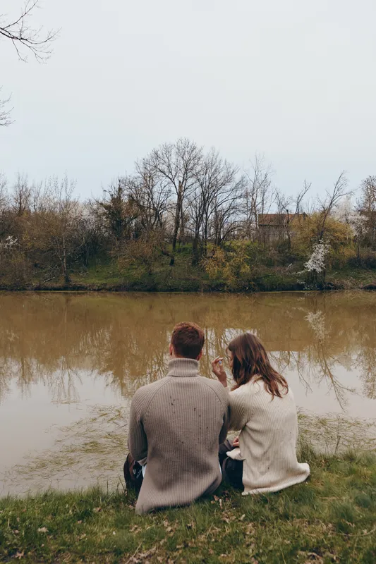 Couple sitting by a calm lake in autumn, bundled in knit sweaters, sharing a quiet moment. Warm tones, soft light, and a gentle breeze create a peaceful mood about connection and nature.