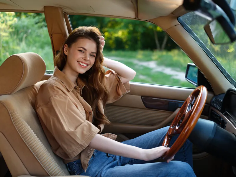 Smiling young woman driving a vintage car, wearing casual beige shirt and denim jeans, joyful summer vibes in a lush green setting.