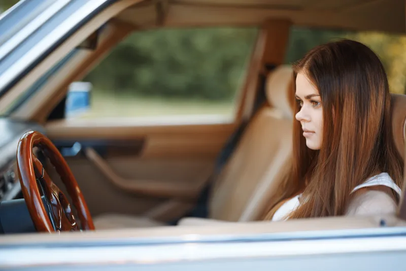 Thoughtful young woman in car interior, deep in contemplation, featuring long brown hair, casual white outfit, natural background, reflecting a serene summer vibe.