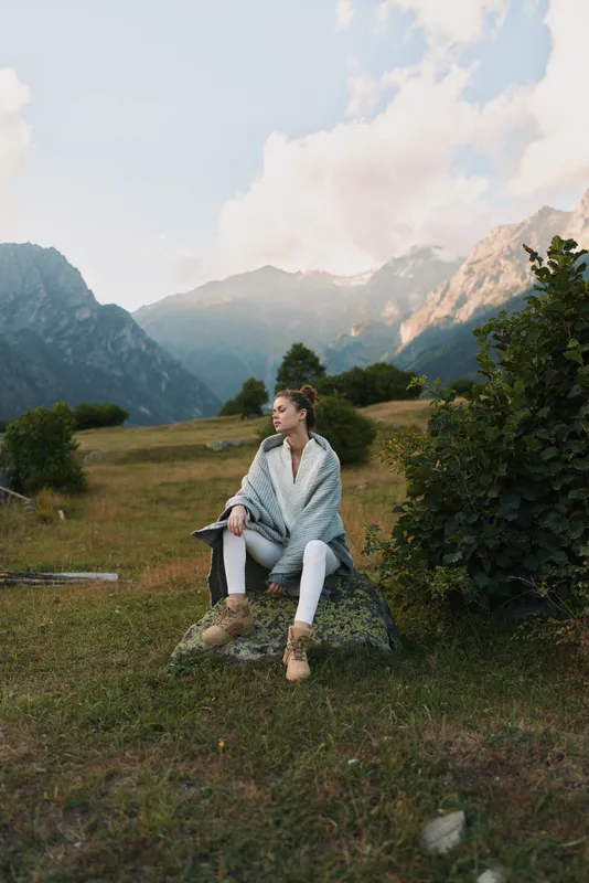A woman sits on a rock in a mountain meadow, wrapped in a soft poncho, enjoying a quiet moment in nature. A serene lifestyle scene of relaxation and exploration.