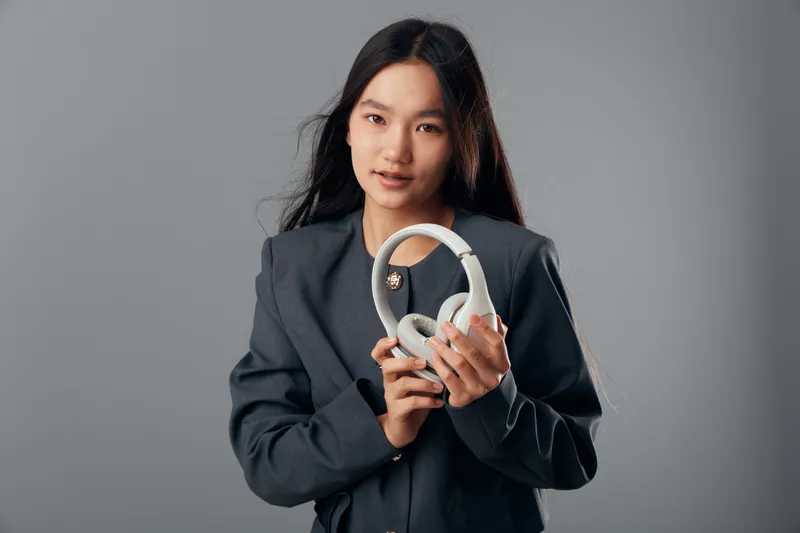 Young woman in dark blazer holding white headphones in hands, posing studio portrait with long hair and natural makeup on gray background with confident expression.