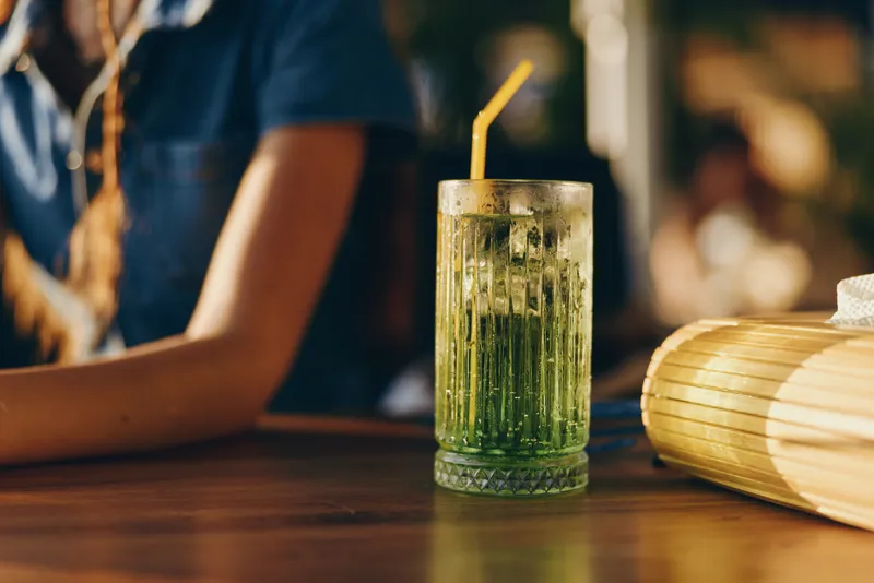 lifestyle woman with modern boho-western denim street style enjoys refreshing green cocktail with straw, film color tones enhance the relaxed urban vibe on wooden table
