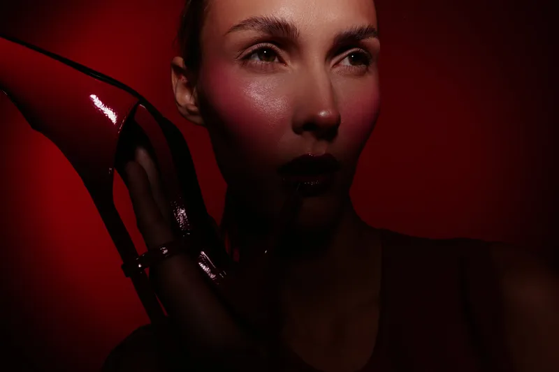 Close-up of a woman with glossy red lips holding a shiny red high heel against a dark red background. Concept of fashion, beauty, and glamour with dramatic lighting.