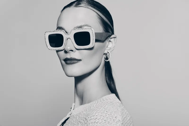 Elegant white woman with pale skin wears large square sunglasses and knitted top, posing confidently in studio lighting.