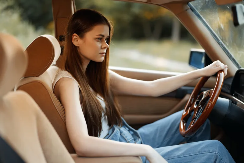 Young woman with long hair driving a vintage car, wearing casual clothing, expressing thoughtful emotions against a serene outdoor backdrop.