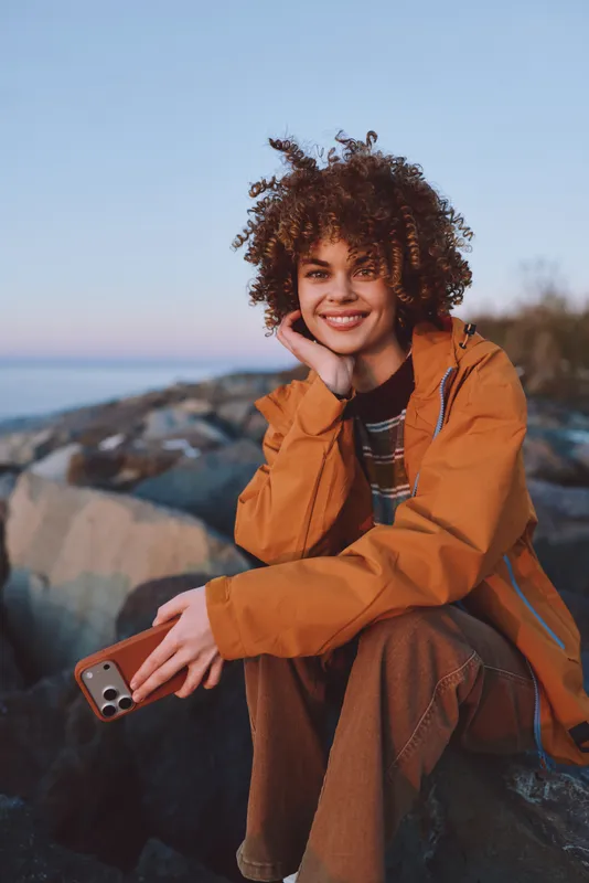 Woman lifestyle portrait outdoors by ocean rocks, smiling, curly hair, wearing an orange jacket, sitting on stone by the shore at sunset, relaxed travel mood, coastal evening