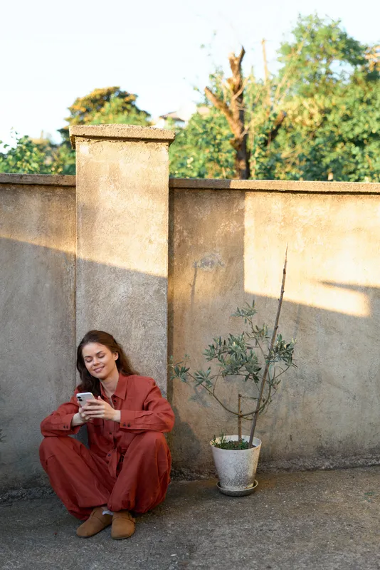 Woman lifestyle outdoor red outfit sitting by wall and using phone beside a plant in a sunlit outdoor scene, candid everyday moment of relaxed posture, the scene radiates calmness and simplicity