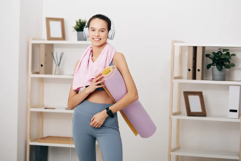 Woman in athletic wear with a yoga mat, headphones, and a bright home studio, ready for a workout and healthy routine.