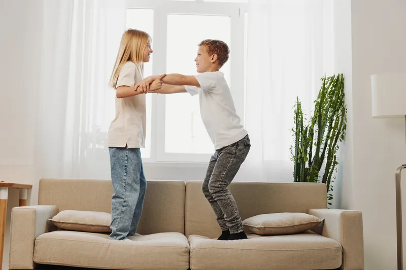 Two children stand on sofa holding hands and smiling in bright living room with modern decor and natural light. Concept of playful childhood and sibling fun at home.