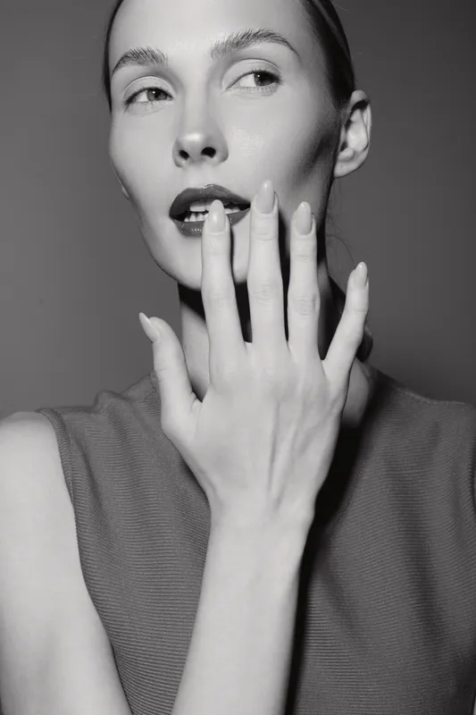 Portrait of a fair skinned Caucasian woman with slicked back hair touching lips, wearing sleeveless top. Studio black and white beauty concept with elegant and mysterious mood.