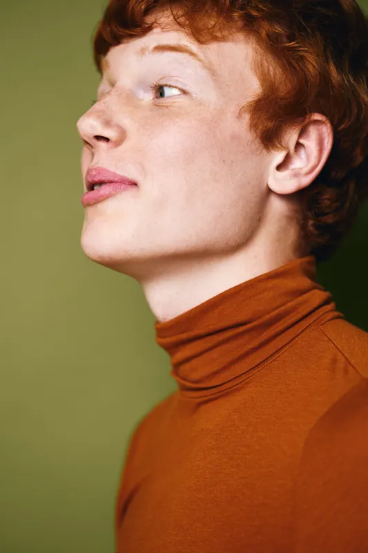Portrait of redhead young man wearing brown turtleneck, looking sideways with thoughtful expression on green background. Profile view, casual fashion and natural light in studio.