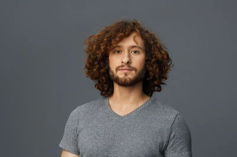 Portrait of a young man with curly hair and a beard, wearing a gray t shirt against a dark background, exuding a calm and confident expression