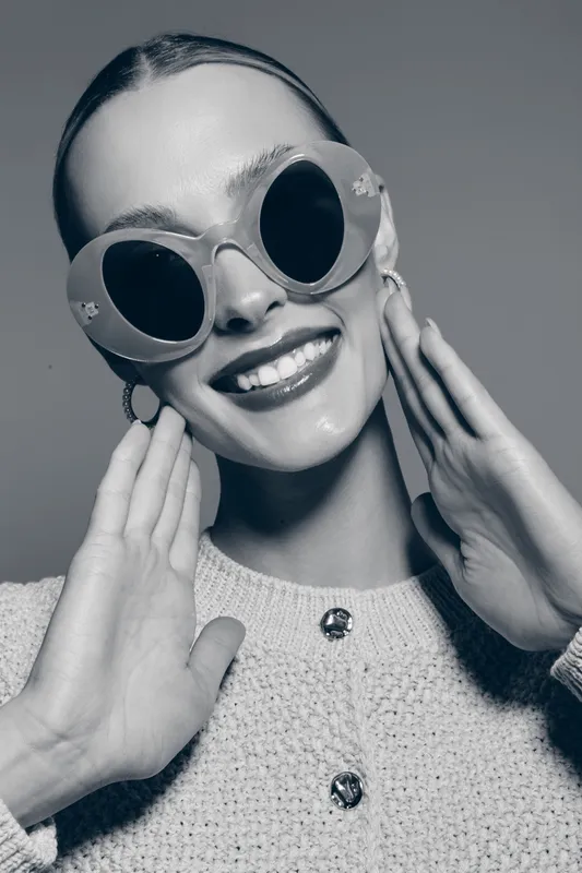 Smiling white woman with fair skin wearing round sunglasses and knit sweater touches face. Fashion portrait with joyful expression on gray background. Concept of style and confidence.