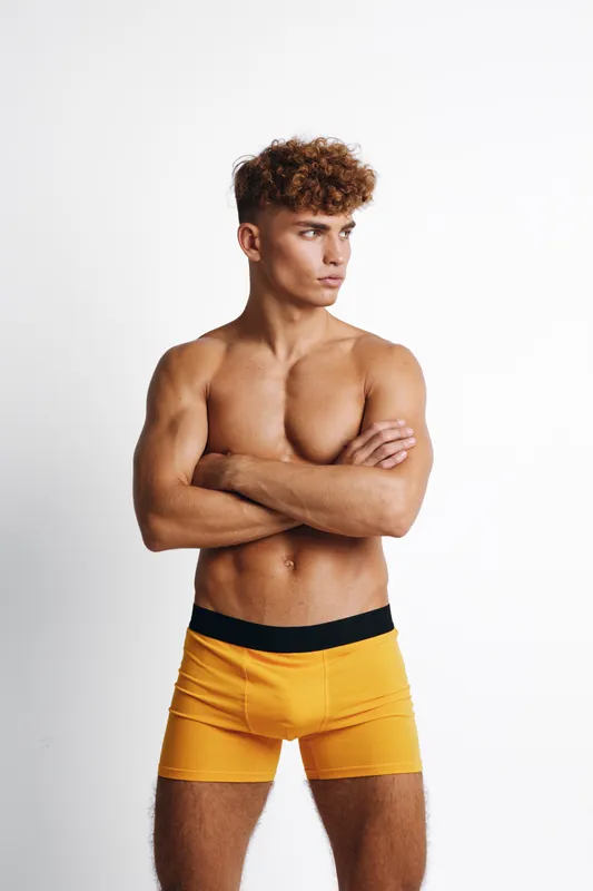 Young muscular man posing shirtless with crossed arms wearing orange shorts, looking to the side on white background. Male fitness, bodybuilder and model portrait.