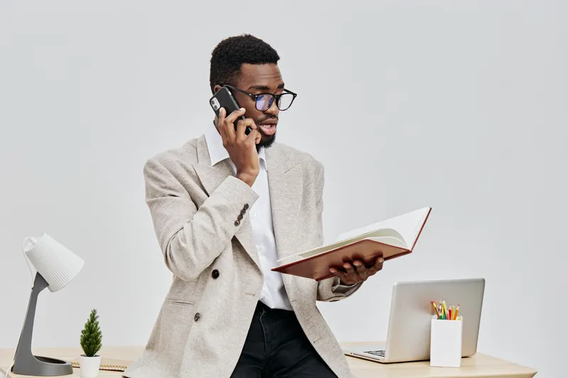 Professional young African American man in smart casual wear talking on mobile phone and reading a notebook, seated at desk with office supplies and laptop, modern work environment. People lifestyle c