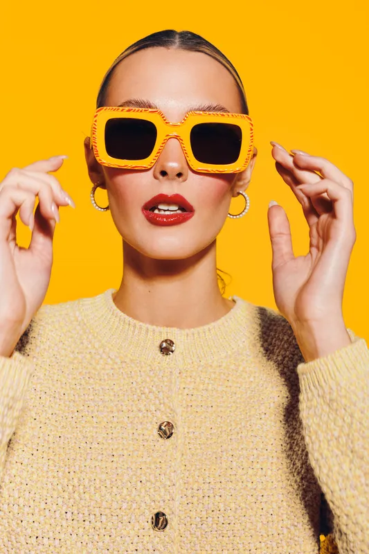 Stylish woman wearing bright orange sunglasses and beige sweater poses against vibrant yellow background. Fashion portrait with bold colors and confident expression for trendy style concept.
