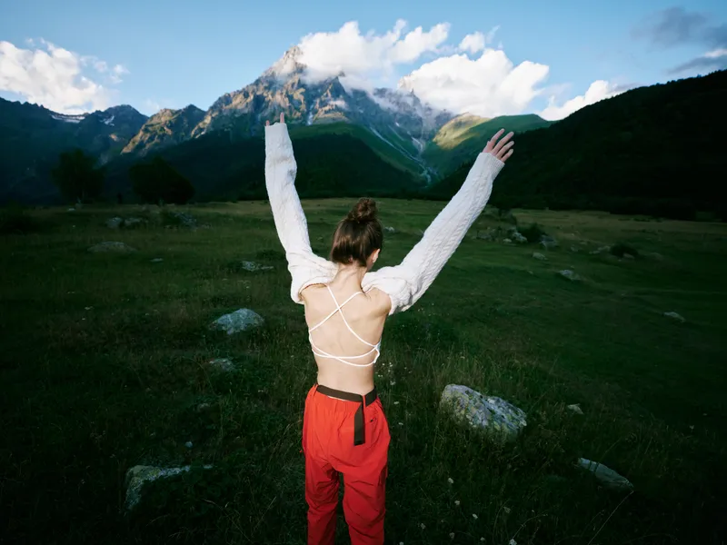 Outdoor scene with mountain landscape, freedom pose, arms raised, open back, green meadow under daylight, wind in trees, nature setting for wellness, adventure and personal growth