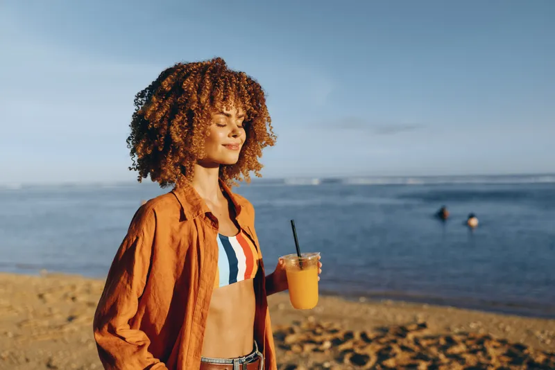 woman beach ocean summer travel relax lifestyle curly hair, standing on sandy shore, holding a glass of orange drink, warm sunlight, distant swimmers, calm coastal mood, outdoor photo