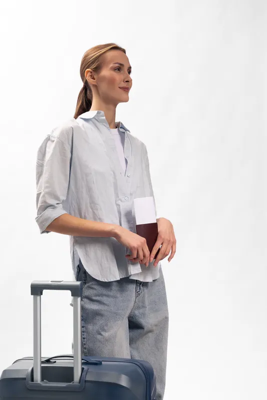 Young woman traveler holding a passport and standing next to a suitcase on a white background. Concept of travel, vacation, and planning a trip with casual clothing and relaxed posture.