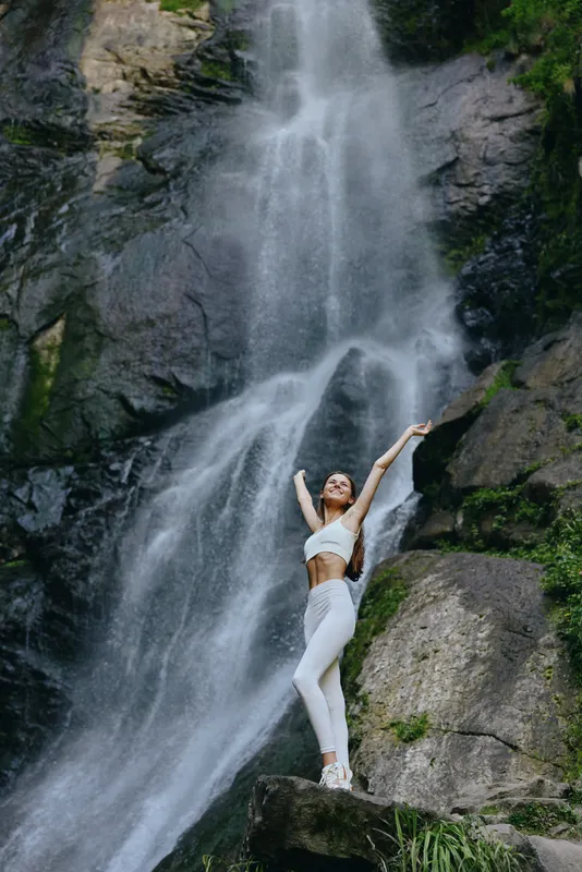 smiling woman in white activewear stretching arms near large waterfall surrounded by lush green rocks and mist, showcasing outdoor fitness and freedom.