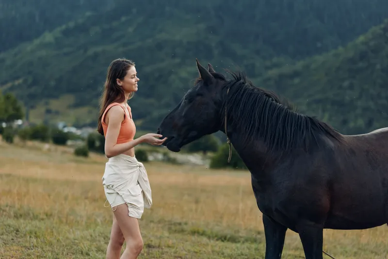 A young girl in a coral top feeds a curious horse in a tranquil meadow with tall grasses and distant mountains, capturing a moment of gentle connection and outdoor serenity.