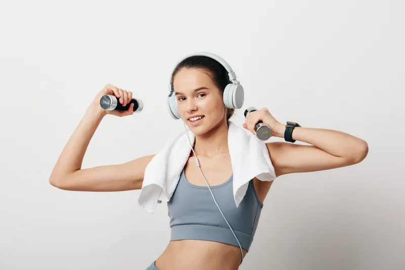 Woman fitness in action: a female athlete lifts dumbbells with headphones on, towel over shoulders, during a high-energy workout in athletic wear at a bright studio.
