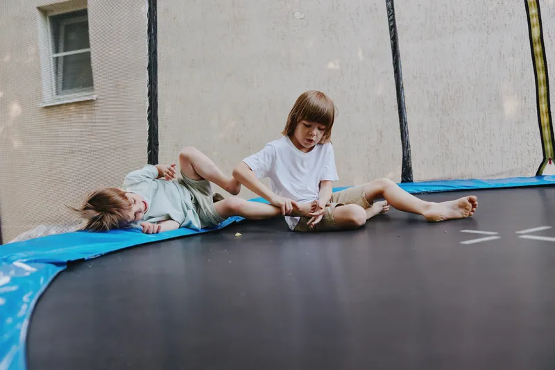 Two playful children, one sitting and the other lying down, enjoy a fun moment on a trampoline. Their laughter fills the air as they bounce joyfully together.