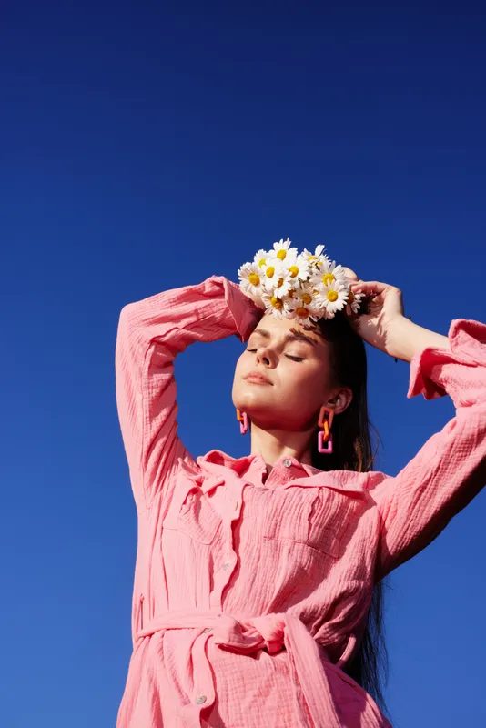 Woman in a pink dress with floral crown against a blue sky, embodying joy and femininity, perfect for seasonal and lifestyle imagery