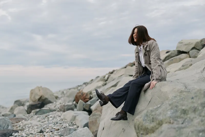 Young woman sits on rocky shore wearing casual clothes, looking away and smiling. Overcast sky creates calm atmosphere by the sea. Concept of relaxation and nature escape.