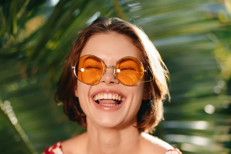 A cheerful woman wearing round orange sunglasses smiles broadly in a bright outdoor setting, surrounded by tropical greenery, creating a vibrant, sunny mood and positive energy.