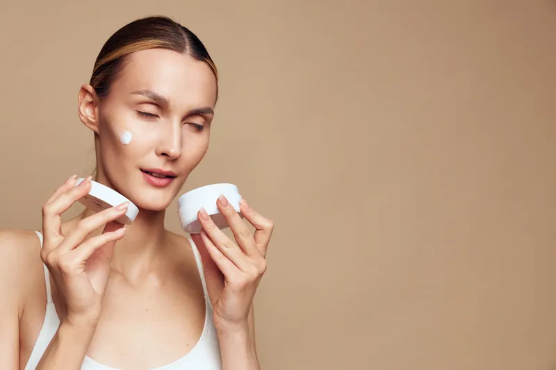 Close up of a young woman applying white face cream on her cheek, with a soft beige background, expressing skincare and self care, calm studio lighting creates a serene mood.