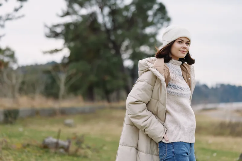Woman standing in a windy field wearing a warm coat, knit sweater and beanie, natural outdoors with trees and grass, casual winter fashion moment