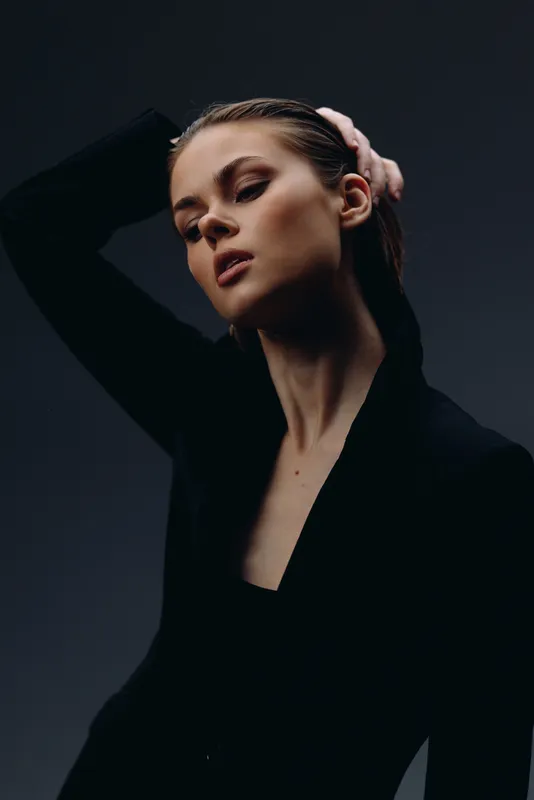 Elegant woman posing with closed eyes against dark background in minimalistic style and soft lighting.